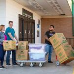 A Moving Helper pushes a dolly with two moving boxes on it. The couple walking behind the helper has the wife carrying a moving box while the husband pushes a dolly with multiple items on it.