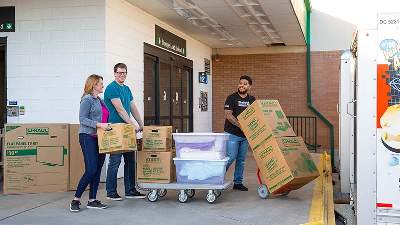 A Moving Helper pushes a dolly with two moving boxes on it. The couple walking behind the helper has the wife carrying a moving box while the husband pushes a dolly with multiple items on it.