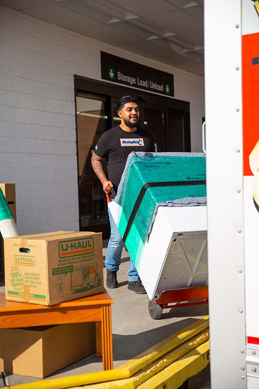 A labor-only mover pushes a dolly carrying a washing machine as he walks toward the U-Haul truck rental.