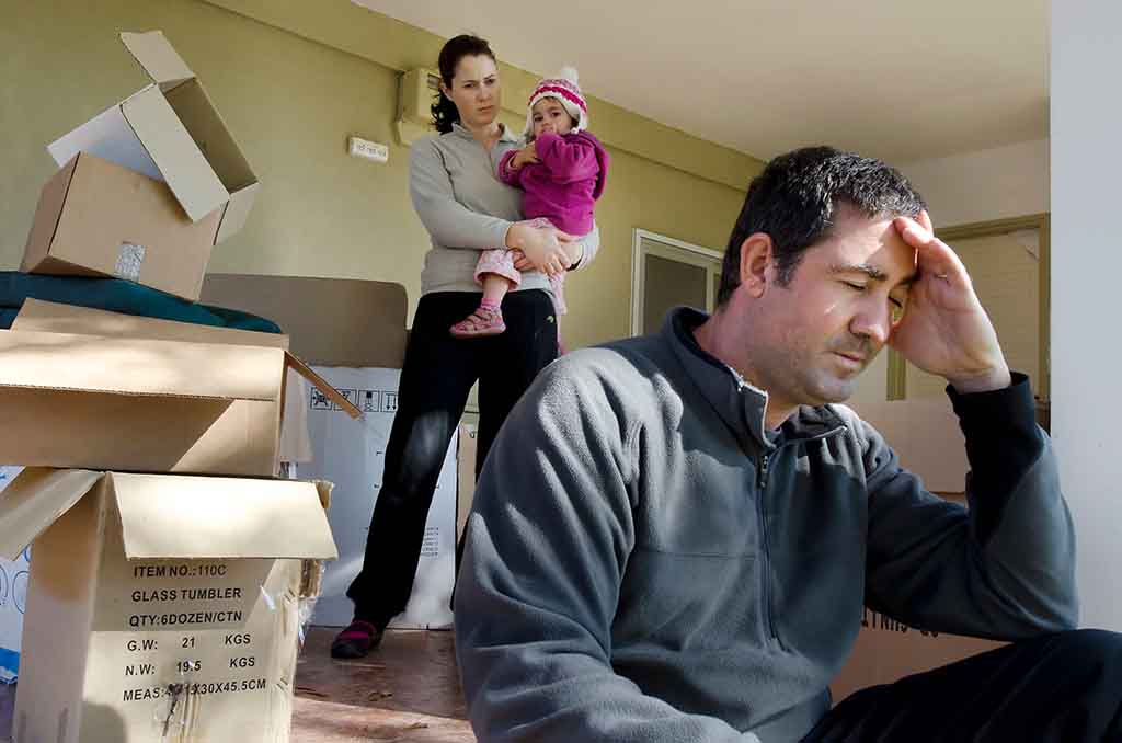 A woman standing holds her daughter as they look at the daughter’s father who is sitting on the ground with his hand pressed against his face. Moving boxes lie around them as they’re in the middle of a divorce move.