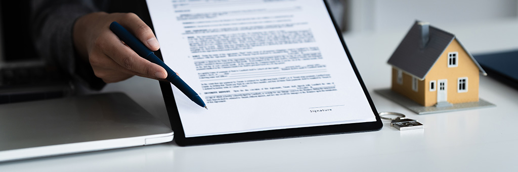 A man holds a pen and a tablet while pointing where a person needs to sign. A computer sits to the left of the tablet, and a key ring and a model house sit to the right of it.