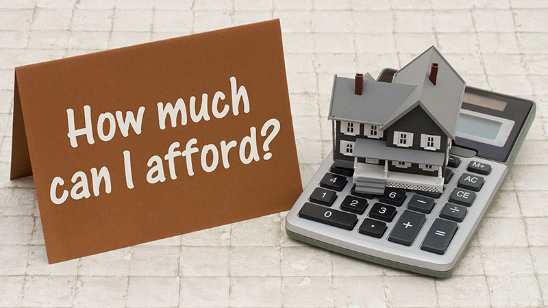 A notecard with the words “How much can I afford?” rests on a table along with a calculator with a model house sitting on top of the calculator, which sits next to the notecard.
