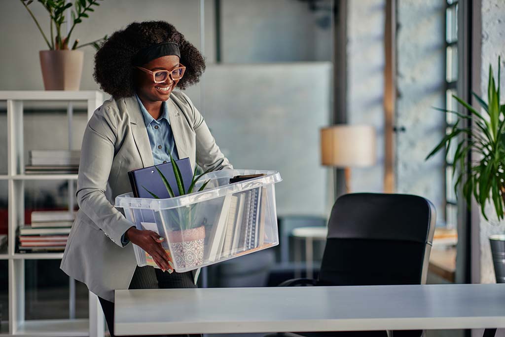A woman begins to set down a box of her office items onto her desk at her new job. Job relocation assistance is possible, but whether someone can receive it will depend on multiple factors.