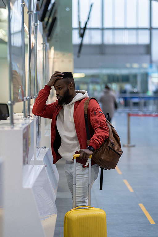 A man put his hand on his head as he looks frustrated while carrying a backpack and holding onto a suitcase at the airport.