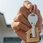 A real estate agent holds keys with a house-shaped keychain in front of a home.