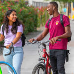 A man and a woman walk together while holding their bicycles when leaving their apartment building. “Should I live alone or with roommates” is an important question to ask when deciding where to live.