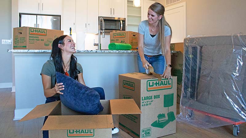 Two women are unpacking their moving boxes in their living room as they prepare to live together as roommates.