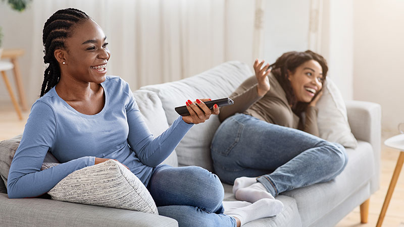 Two women smile and laugh while sitting on the couch together. One of the women is holding a TV remote to turn up the volume while watching a show.