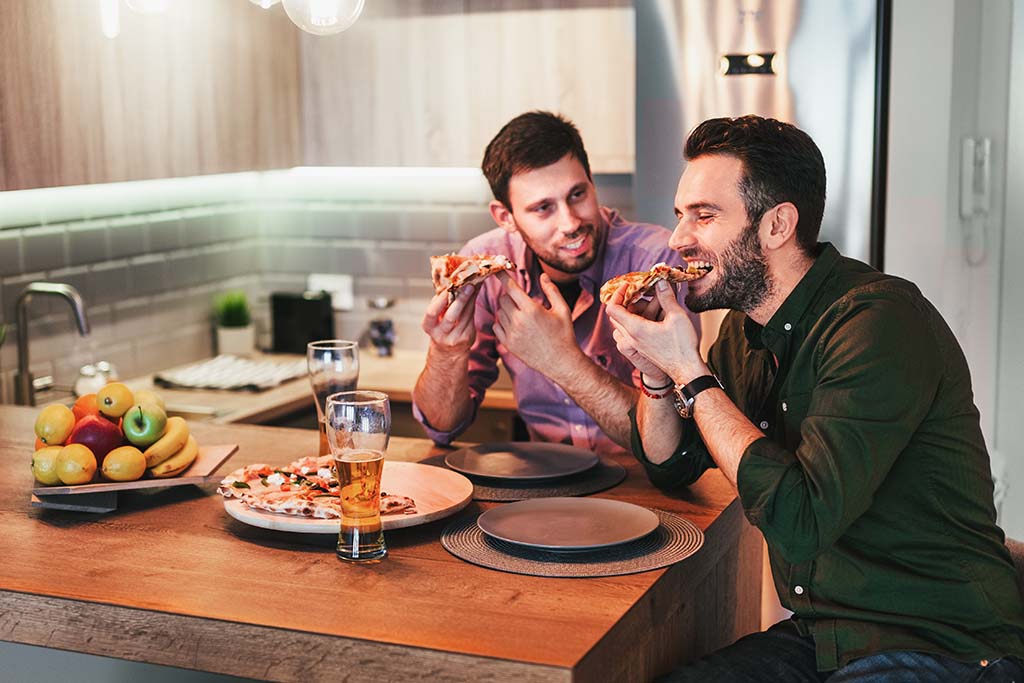 Two male roommates eat pizza together on the bar countertop area in their kitchen. “Should I live alone or with roommates” is an important question to ask when deciding where to live.
