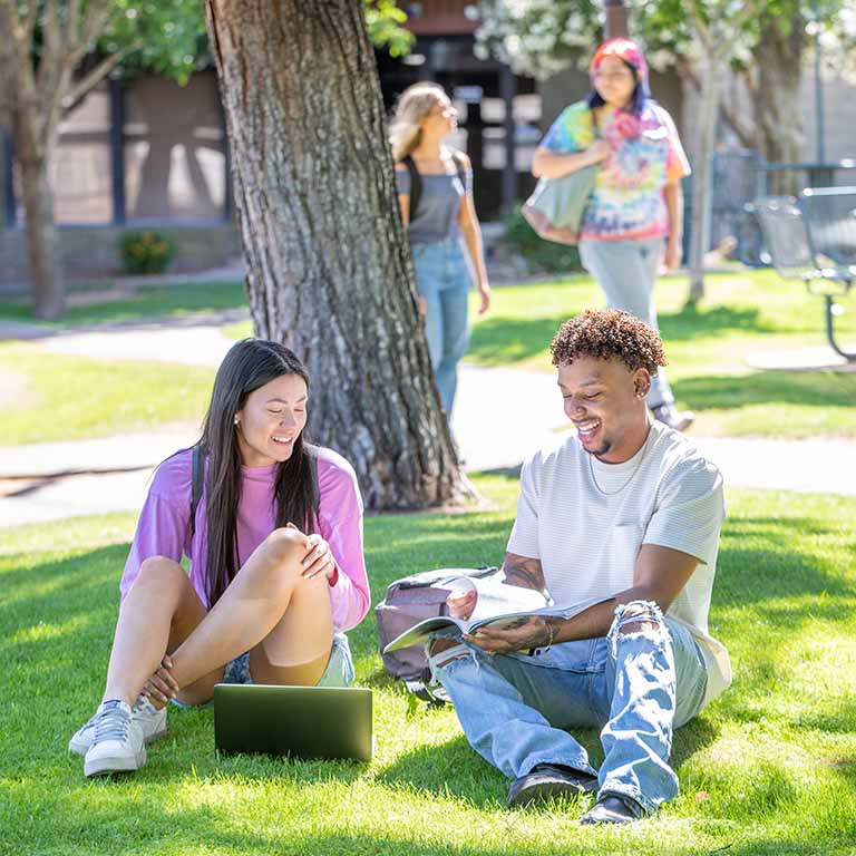 A male and female roommate sit on the grass outside underneath a tree’s shade in their apartment complex’s common area. “Should I live alone or with roommates” is an important question to ask when deciding where to live.