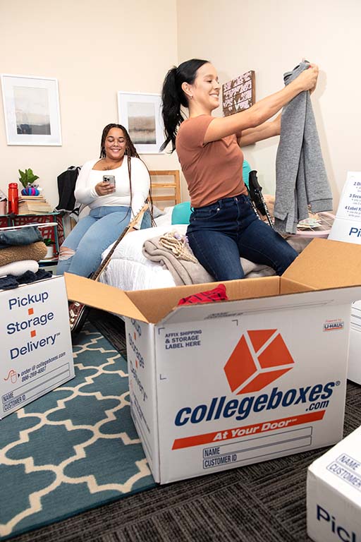 A female freshman packs her clothes into a Collegebox while her roommate looks at her phone in the background.