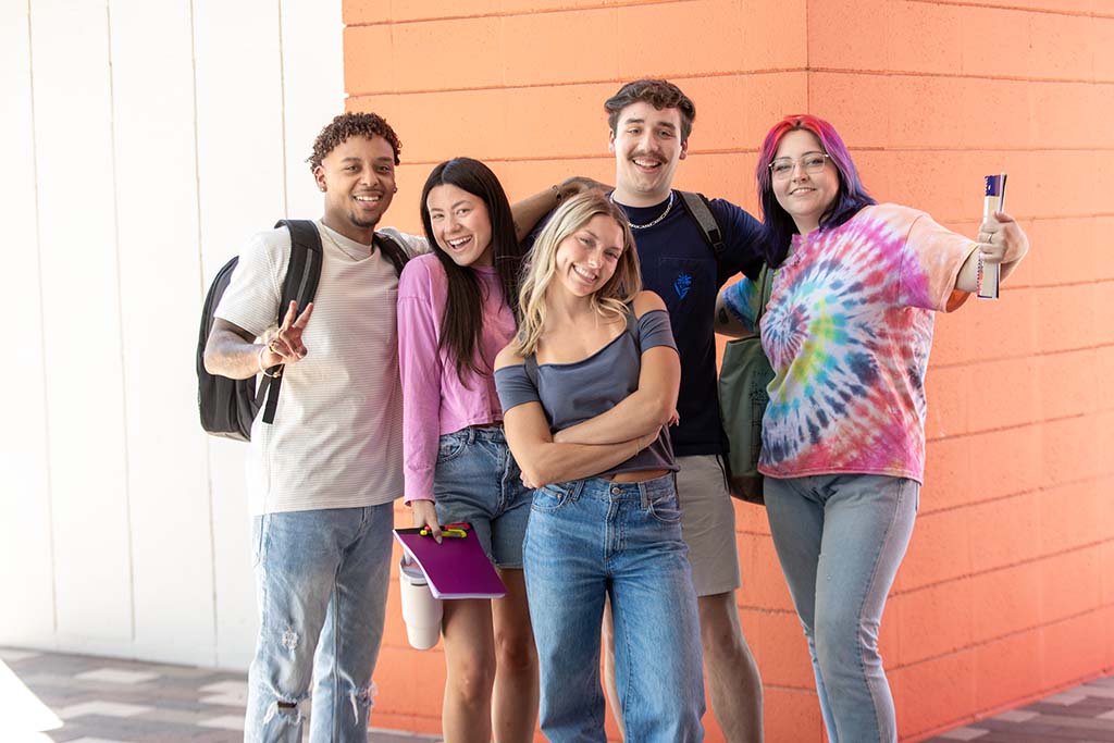 A group of freshmen — three females and two males — stand together as they smile for a group photo on campus.