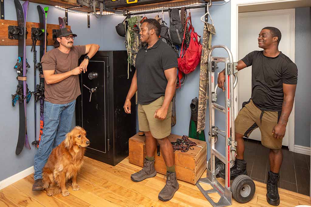 A man and his dog stand next to their gun safe while talking to two Moving Help Service Providers who will prepare to move the gun safe.