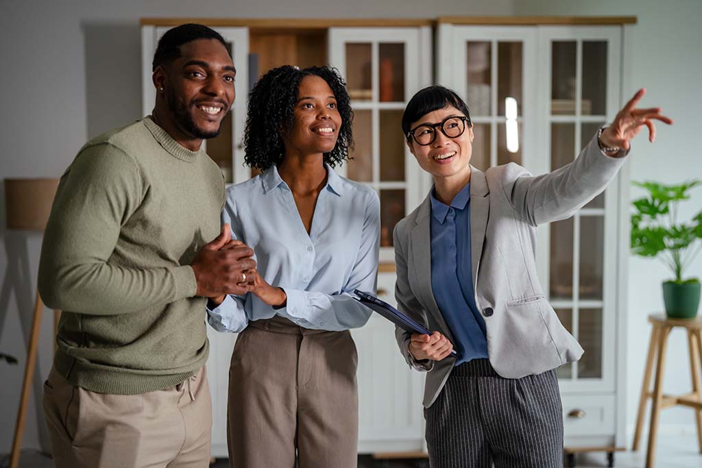 A husband and wife look toward the area of a house the female real estate agent is pointing at for this first-time homebuyer couple.