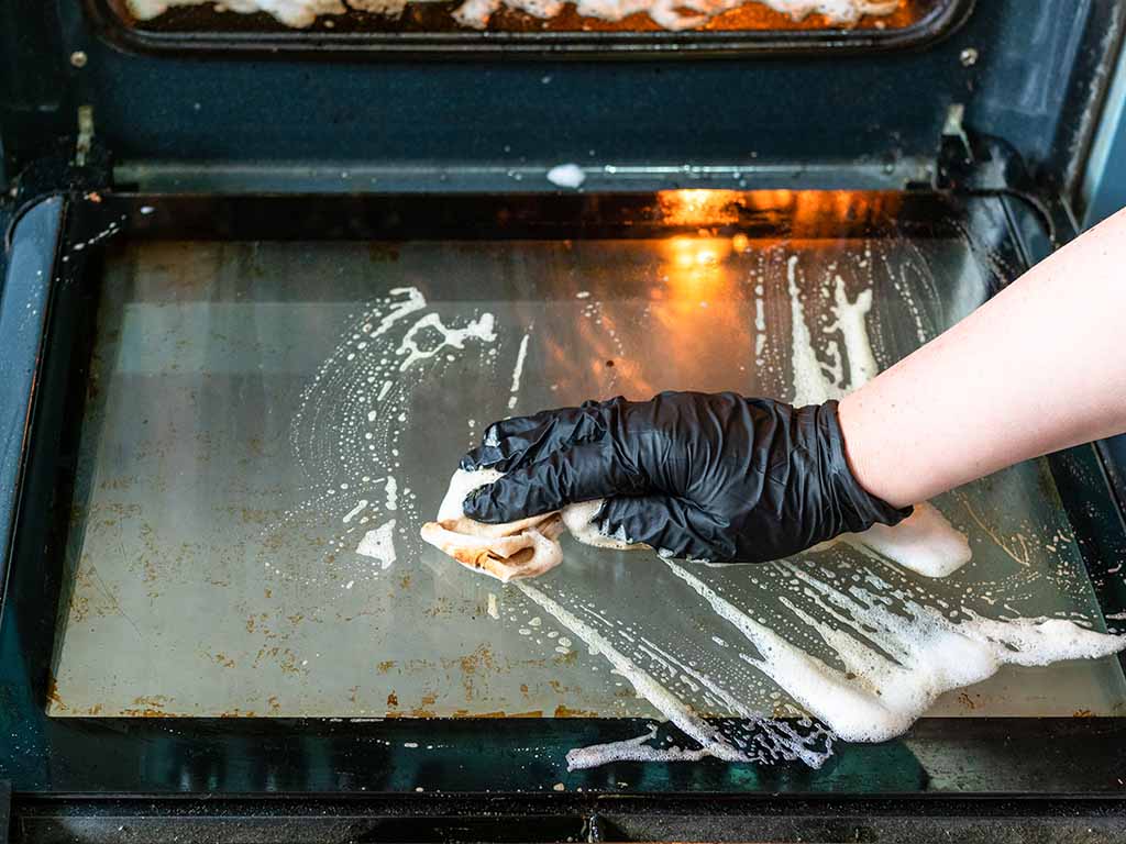 A woman cleans the oven’s glass door with soap and a dish cloth.