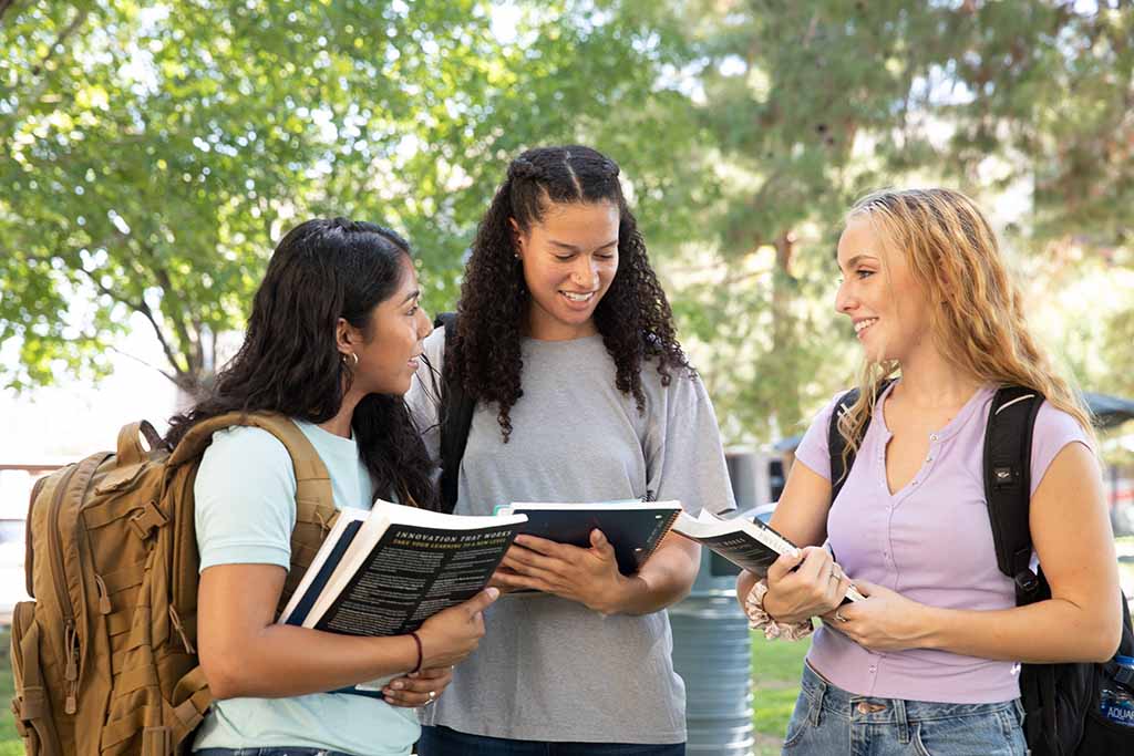 Three college women stand together while talking to one another. The college students carry backpacks and textbooks.