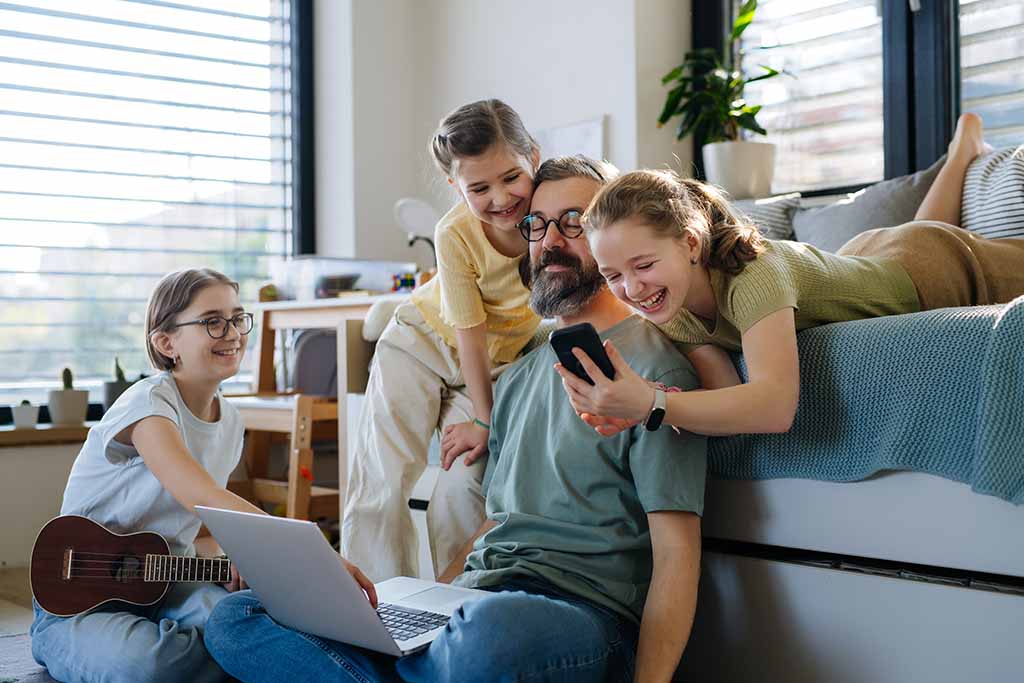 A man works on his computer while sitting on the living room floor. The man’s three daughters surround him on the couch and on the floor.