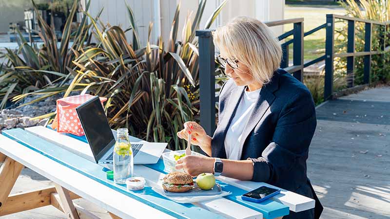 A woman sits outside to eat her lunch while working on her computer through her lunch break.