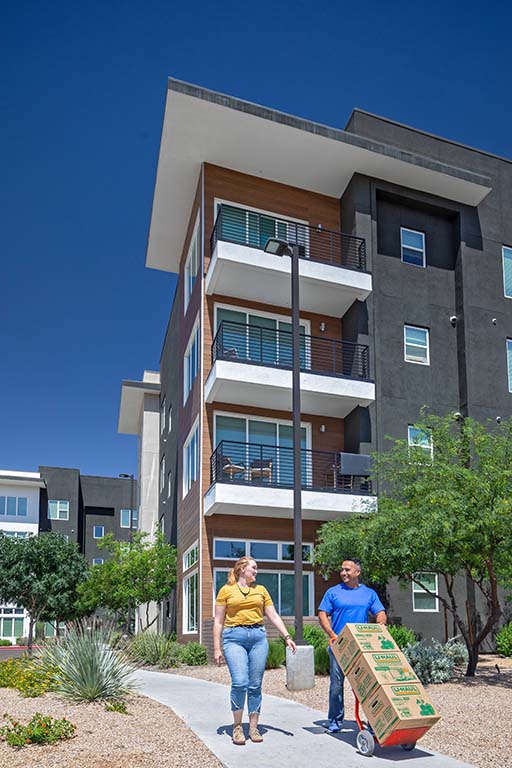 A man and a woman walk outside an apartment, and the man is pushing boxes. The woman is moving into a new apartment after completing her apartment search.