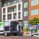 Two men carry a couch out of a truck and into an apartment building. It’s important to consider the pros and cons for an apartment vs. a condo.