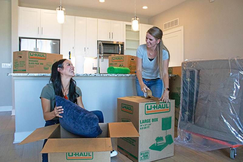 Two women unpack boxes in a new apartment. When deciding to try to live in an apartment vs. a condo, you should consider the pros and cons for each housing type.
