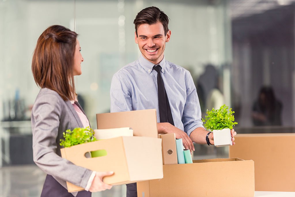 A male and female co-worker smile at each other while packing a desk. The female co-worker is leaving for a new job where she got employer relocation assistance.