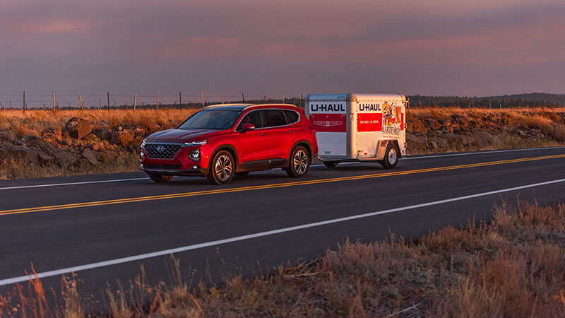 A car pulls a U-Haul cargo trailer behind it on a highway while the sun begins to set.