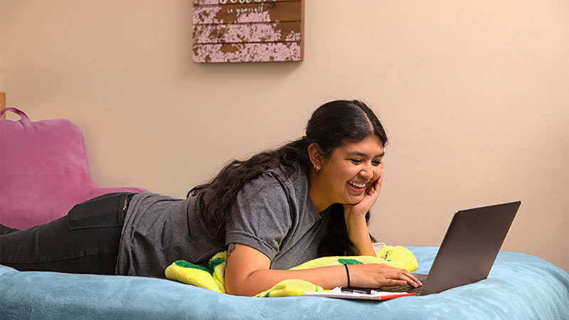 A girl lies on her bed, laughing while looking at her laptop. She’s living alone in her apartment.