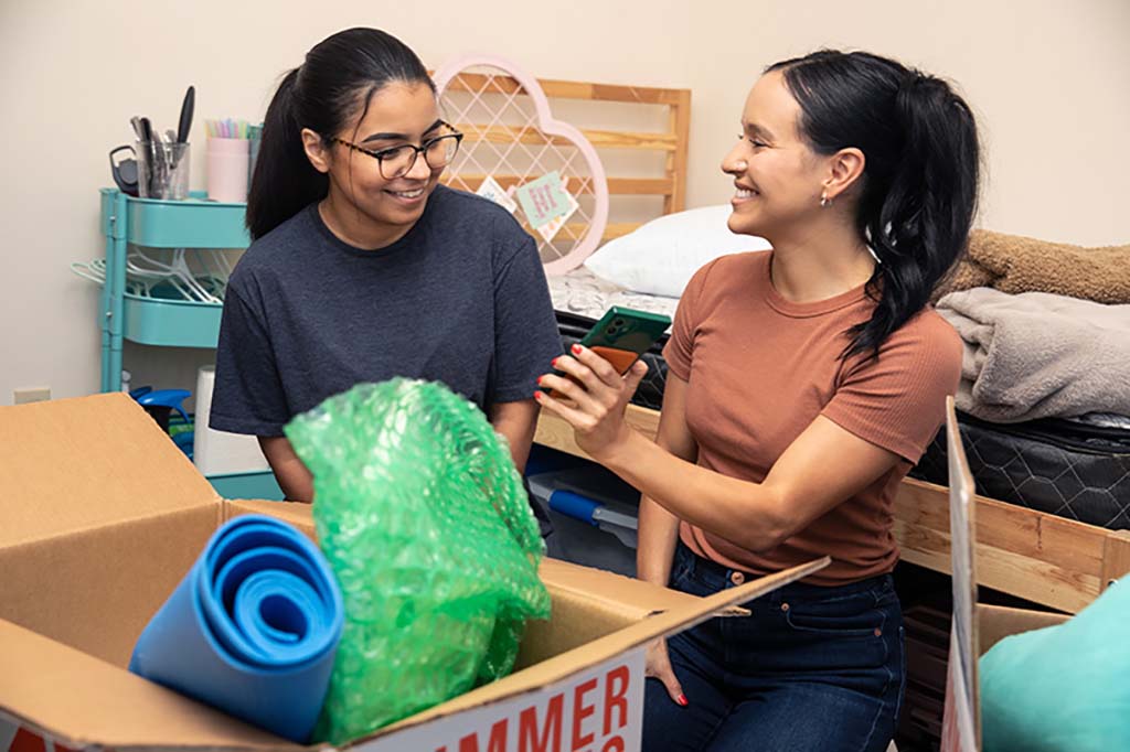 Two girls sit in their room, and one girl is showing the other something on her phone. They’re in the process of unpacking boxes together because they’re roommates.