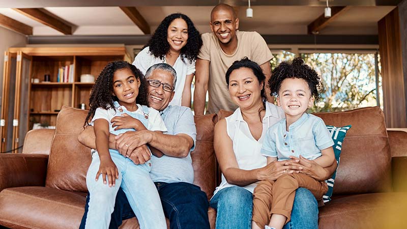 A multigenerational Canadian family, including grandparents, a mother, a father, and their children, relaxes together on a living room couch.