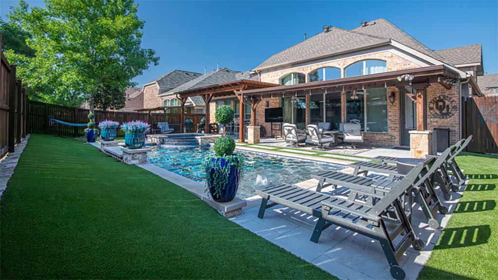 The backyard of a house contains a pool, lawn chairs, and potted plants surrounded by artificial turf.