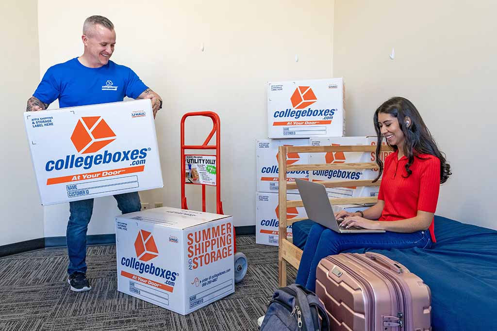 A woman is on her laptop in her college dorm. A Service Provider is bringing Collegeboxes into the room.