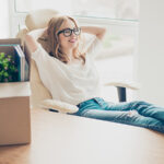 A woman smiles while relaxing in her chair as a moving box sits on the desk in her office as she prepares for her new job. It’s easy to learn “What is a relocation allowance?”