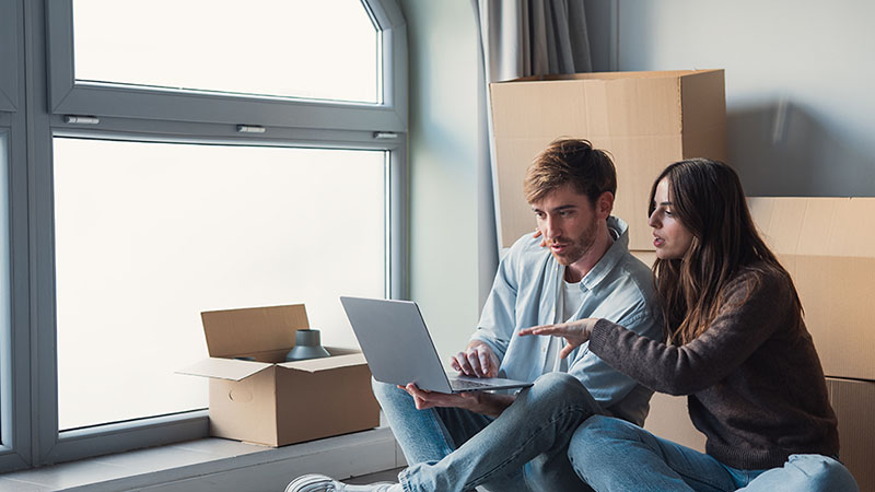 A man and woman sit on the ground while looking and pointing at the laptop the man is holding as moving boxes surround the couple before they have a job relocation move.