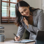 A woman writes something down on a notebook while talking on the phone and looking at her tablet in her office. The woman is smiling because she got the relocation package she wanted for her new job.