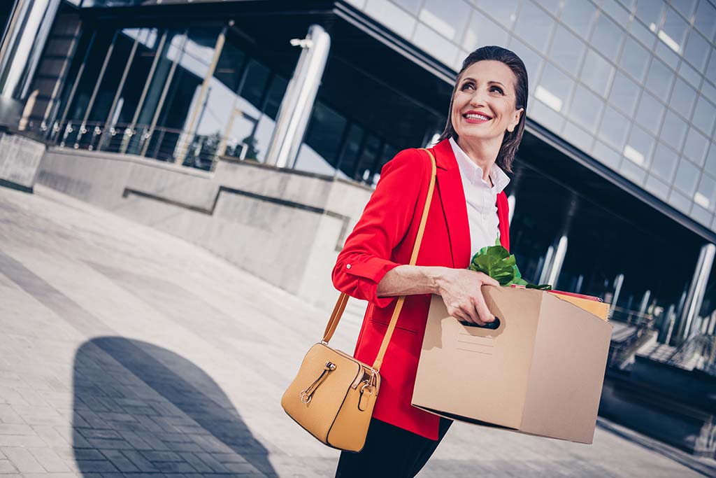 A woman smiles while carrying a box of her office items as she leaves her old job’s office buildings to start preparing for her job relocation.
