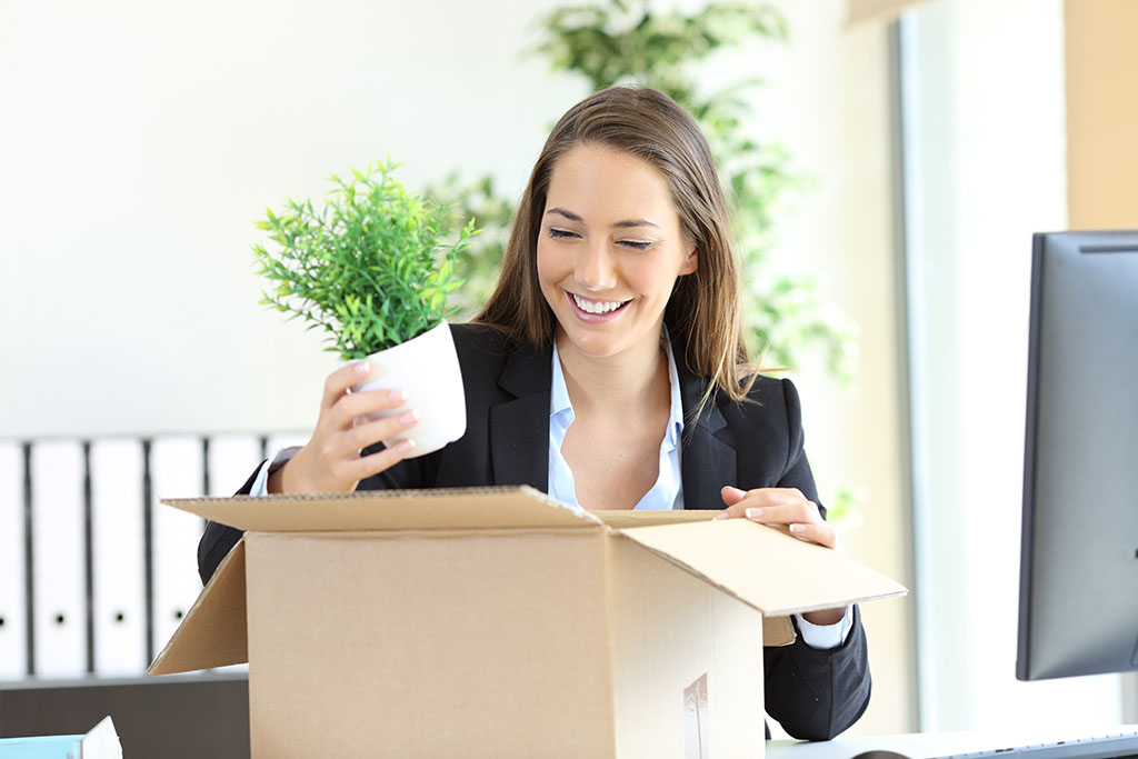 A woman smiles while she packs her office plant into her moving box. The woman is leaving her old job for a new job after receiving the relocation package she wanted for her new job relocation.