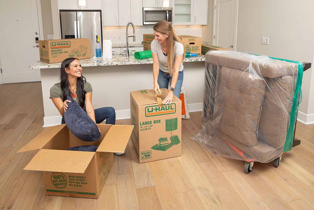 Two women unpack boxes in their new apartment. They’re sharing a kitchen and a living space in their apartment.