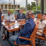 A woman is standing, and a group of people is sitting in a conference room in an office building. When moving for work, relocation packages offer many moving benefits.