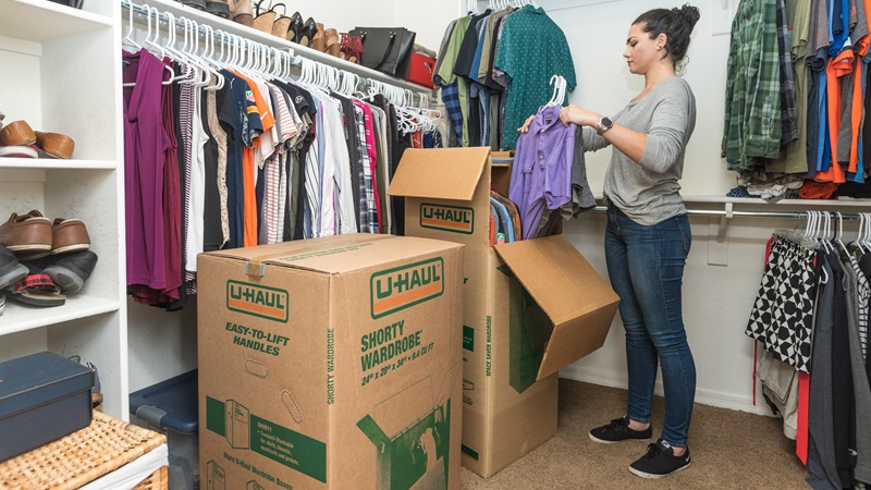 A woman is moving hanging clothes from her closet into a shorty wardrobe box.