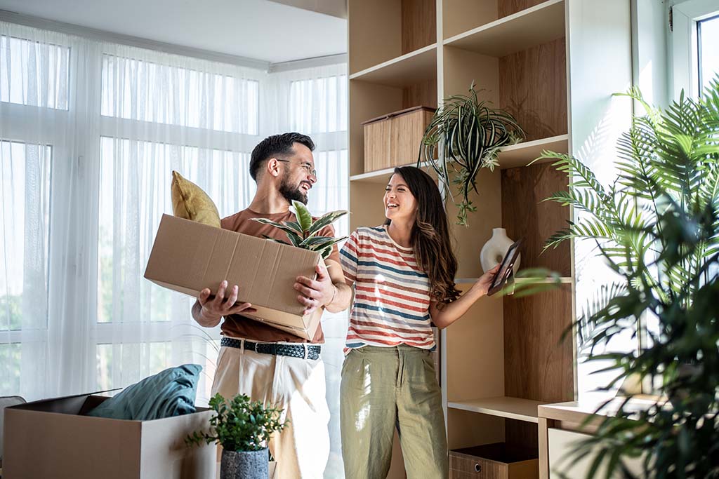 A couple works together to pack the plants in their living into a moving box the husband is carrying while the wife grabs the plants.