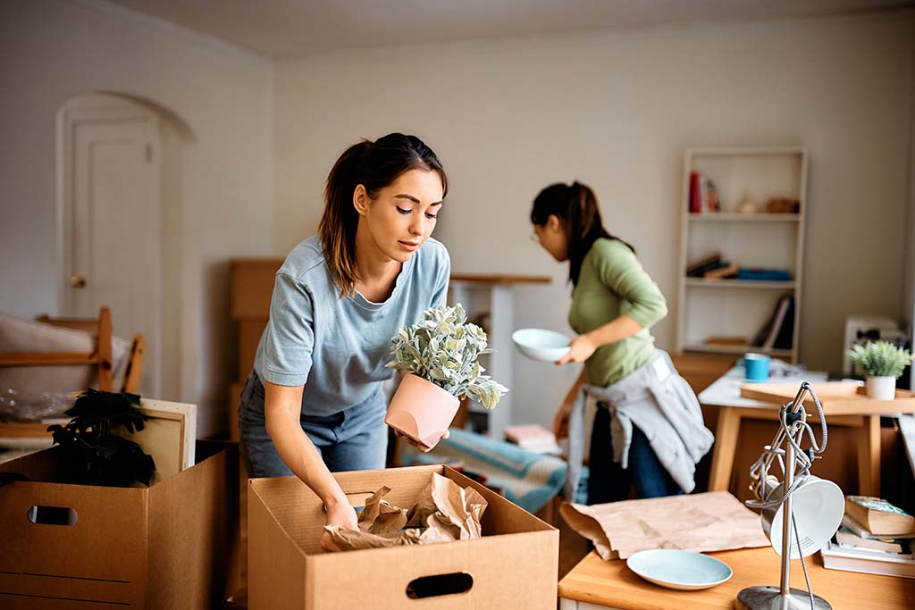 A lesbian couple unpacks plants out of their moving boxes and place them inside the living room of their new home.