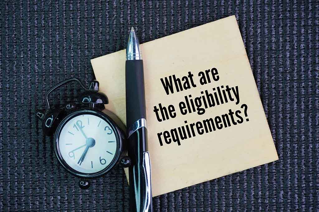 A piece of paper has the words “What are the eligibility requirements?” written on it. A pen and an old-fashioned bell alarm clock sit to the left and partially on the note.