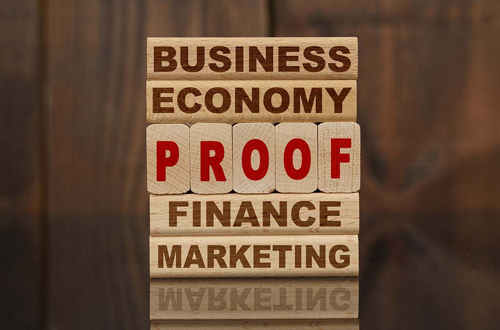 Five blocks sit on top of each other vertically on a table with a reflection. Each block has a different word written on it. The five words are: “Business,” “Economy,” “Proof,” “Finance,” and “Marketing.”