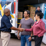A Moving Helper shakes hands with the couple as two Moving Helpers load the couple’s furniture into a U-Haul truck rental in the background.