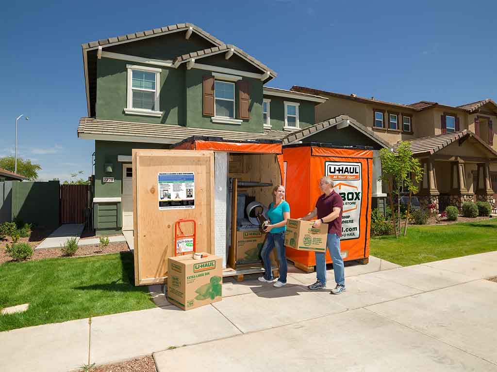 A couple loads their U-Box storage containers with moving boxes. The U-Box containers sit in front of the couple’s house.
