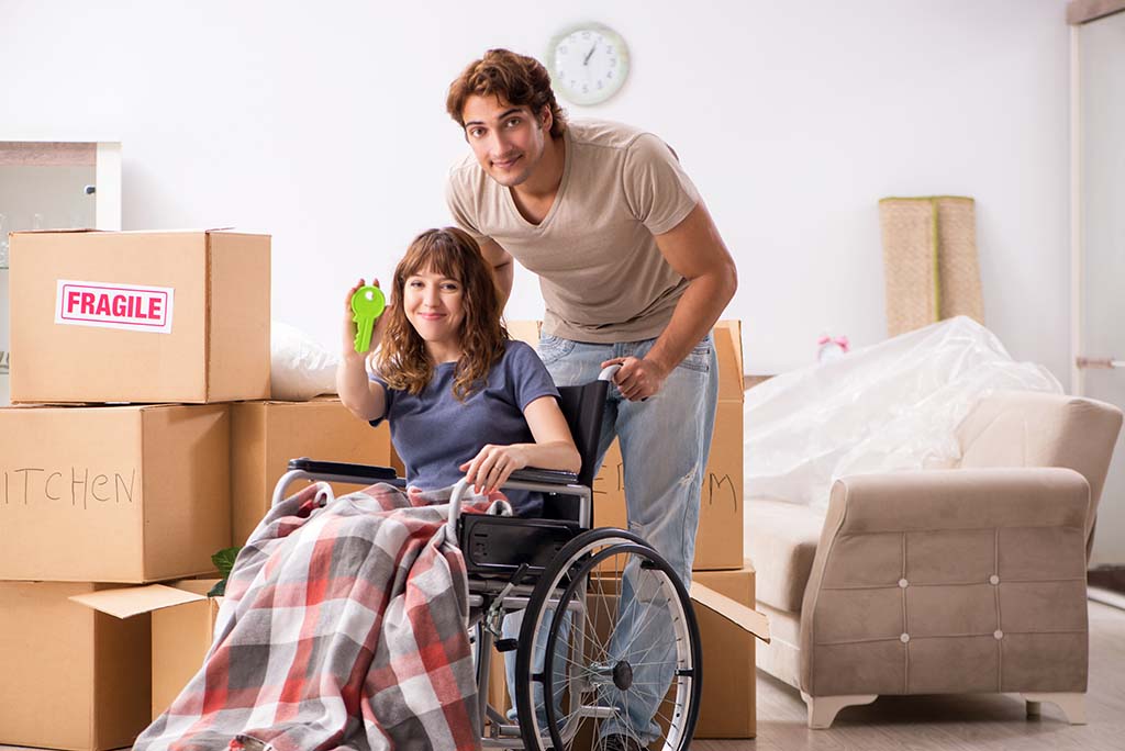 A man stands behind his girlfriend in a wheelchair as they smile together about getting the keys to their new home.