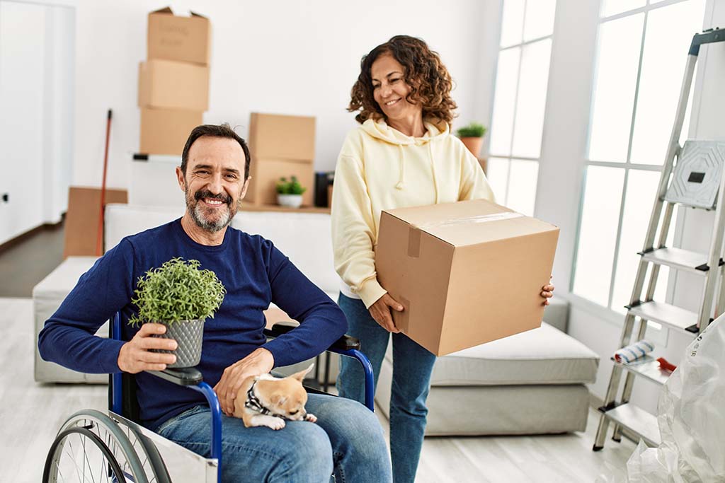 A man sitting in a wheelchair smiles as his wife carries a moving box next to him.