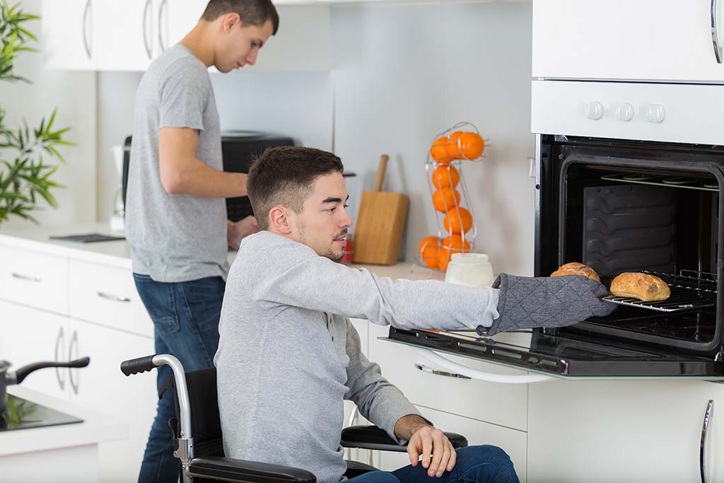 A gay couple cook together in a kitchen. One of the men sits in a wheelchair while using oven mittens to pull bread out of the oven while the other man chops vegetables on the kitchen countertop behind him.