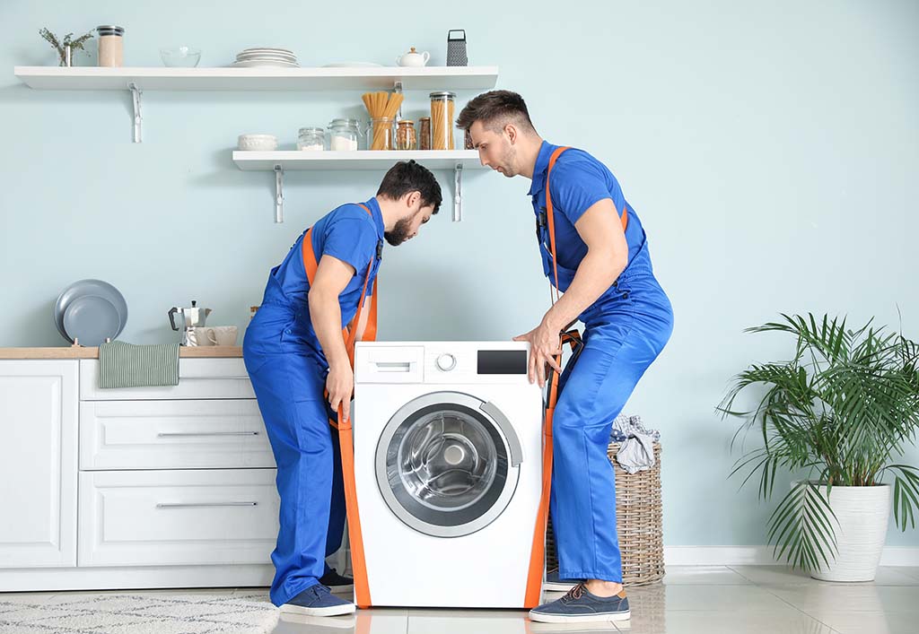 Two movers carry a washing machine with straps on their shoulders.